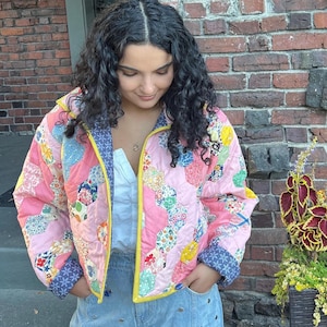 May include: A quilted jacket featuring a pink hexagonal patchwork pattern with various floral prints, a bright yellow trim along the zipper, and dark blue patterned cuffs. The jacket is worn over a white collared shirt and light blue denim jeans.