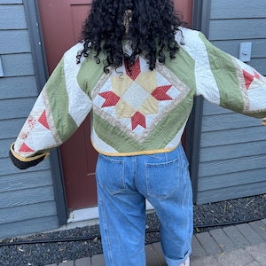 May include: A cropped quilted jacket featuring a central star pattern in cream, red, and tan, with olive green and white panels. The jacket has long sleeves with red and white floral accents, a yellow hem, and is paired with blue denim jeans.