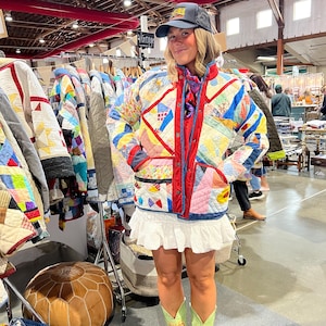 May include: A multi-colored patchwork quilted jacket with red trim, a white ruffled mini skirt, and light green cowboy boots with red embroidery. The outfit is paired with a dark grey baseball cap featuring a colorful graphic patch on the front.