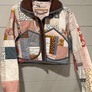 May include: A cropped patchwork jacket with a brown corduroy collar and trim. The jacket features a variety of fabric patterns in shades of pink, blue, white, and gold. It is hanging on a wooden hanger.