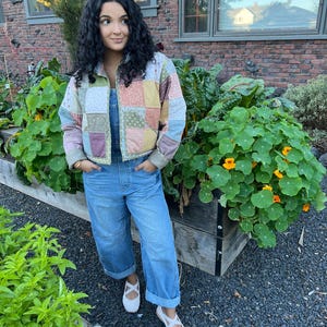 May include: A quilted patchwork jacket featuring various muted floral and geometric patterns in shades of pink, blue, green, and cream, worn over light blue denim overalls with cuffed hems and white flat shoes with a single strap across the top.
