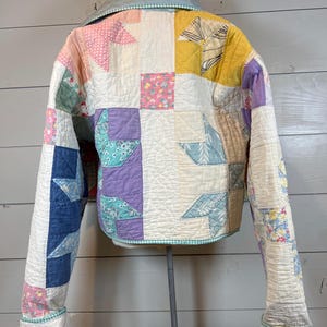 May include: A long-sleeved jacket constructed from a patchwork quilt featuring various fabric squares in shades of yellow, purple, blue, pink, and white. The garment includes a light blue collar and a green and white checkered trim along the edges.