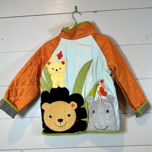 Peut inclure: Une veste matelassée avec des manches orange et un panneau central bleu clair orné d'appliques en tissu représentant un lion jaune, une tête de lion à crinière noire, un hippopotame gris et un petit oiseau rouge sur des feuilles vertes.