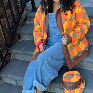 May include: A quilted jacket featuring a geometric pattern in orange, yellow, and brown with a brown trim, paired with blue denim overalls and a matching quilted bucket hat. The outfit is completed with light-colored sneakers with crisscross straps.