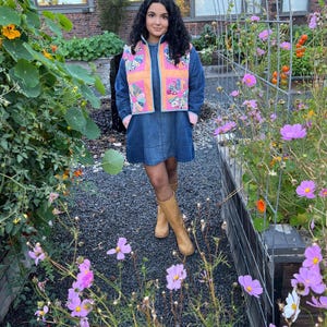 May include: A dark blue denim dress with long sleeves and a high neckline, paired with a quilted vest featuring orange panels and colorful floral fabric squares. The outfit is completed with tan knee-high boots.