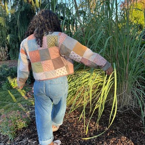 May include: A cropped jacket constructed from a patchwork of quilted fabric squares in muted tones of sage green, dusty rose, mustard yellow, and light blue, paired with light-wash blue denim jeans featuring a straight-leg fit and cuffed hems.