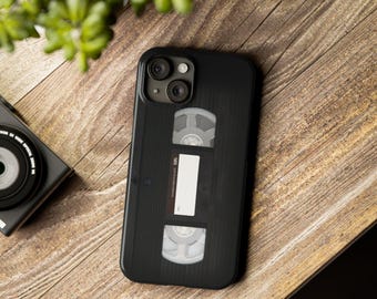 Może przedstawiać: Czarne etui na iPhone z vintage'owym wzorem kasety VHS. Etui ma czarne tło z białą etykietą z napisem "VHS" i białym wzorem kasety.