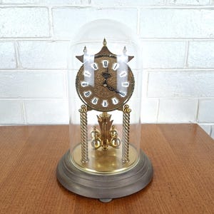 Puede incluir: Un reloj de aniversario dorado con esfera circular, números romanos y péndulo giratorio, encerrado en una cúpula de cristal transparente. El reloj cuenta con dos pilares de latón trenzado y se asienta sobre una base metálica oscura y circular.