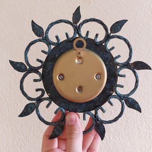 May include: A vintage metal wall decor piece with a circular design. The piece features a dark teal outer frame with leaf accents and a gold-colored center. The back has a gold-colored plate with mounting hardware.