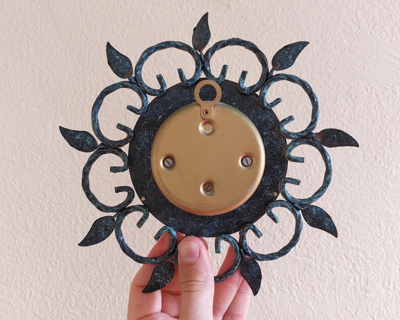 May include: A vintage metal wall decor piece with a circular design. The piece features a dark teal outer frame with leaf accents and a gold-colored center. The back has a gold-colored plate with mounting hardware.