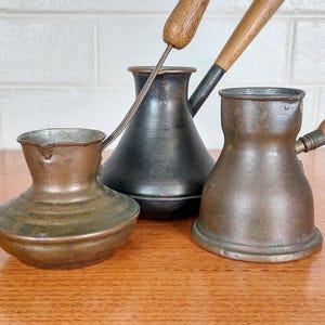 May include: Three antique copper and metal coffee pots with wooden handles. The pots vary in size and shape, with one having a long, thin handle and another a short, rounded handle. The copper has a warm, aged patina, and the metal is dark.