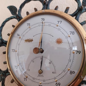 May include: Antique brass barometer with a white face, featuring a needle and a smaller dial. The face has numbers and weather symbols, surrounded by a decorative, dark green metal frame. The barometer measures atmospheric pressure.