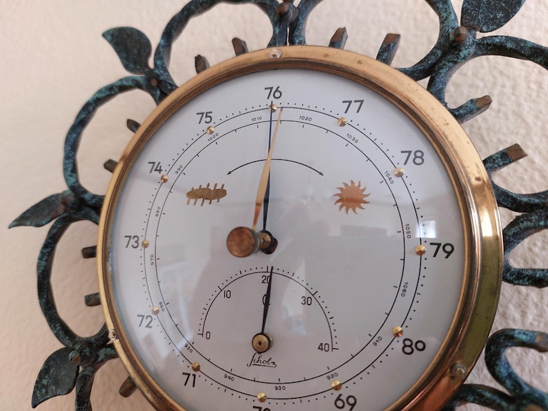 May include: Antique brass barometer with a white face, featuring a needle and a smaller dial. The face has numbers and weather symbols, surrounded by a decorative, dark green metal frame. The barometer measures atmospheric pressure.
