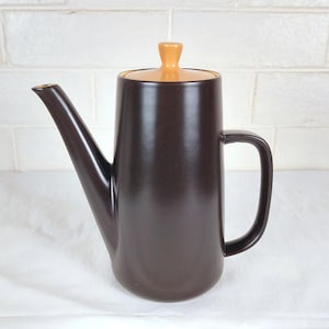 May include: A brown ceramic coffee pot with a long spout and a handle. The lid is a light orange colour. The coffee pot is on a white surface.