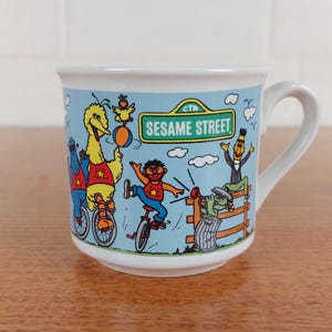 Op de afbeelding: Witte keramische mok met een Sesame Street-ontwerp. De mok heeft illustraties van Big Bird, Elmo en andere personages tegen een lichtblauwe achtergrond. De mok heeft een wit handvat en de tekst "Sesame Street" op de zijkant.