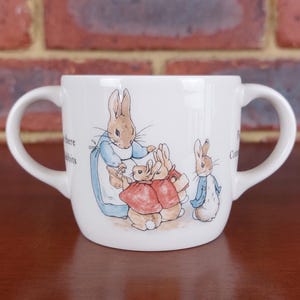 以下が含まれることがあります： 2つのハンドルが付いた白いセラミックマグ。ピーターラビットと他のウサギのカラフルなイラストが特徴です。イラストにはテキストが含まれており、レンガの背景に配置されています。マグは持ちやすいように設計されています。