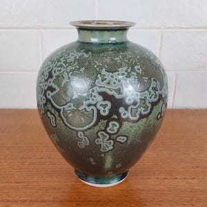 May include: A ceramic vase with a round body and a narrow neck. The vase has a mottled pattern in green, brown, and white. The base has a white rim. The vase is on a wooden surface. The vase is approximately 20 cm tall.