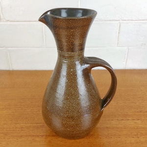 May include: A brown ceramic pitcher with a handle and a spout. The pitcher has a speckled texture and a dark brown interior. It is sitting on a wooden surface.