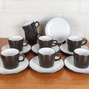 May include: A set of eight espresso cups and saucers. The cups are dark brown with white interiors and black handles. The saucers are white with a black trim. A large white plate with a black trim is also included.