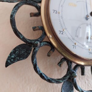 May include: Close-up of a vintage barometer with a white face and gold-colored trim. The barometer displays temperature readings and is set in a decorative, dark green metal frame with leaf and swirl details.