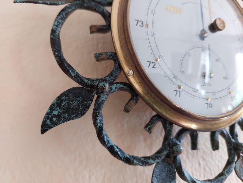 May include: Close-up of a vintage barometer with a white face and gold-colored trim. The barometer displays temperature readings and is set in a decorative, dark green metal frame with leaf and swirl details.