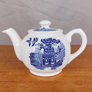 以下が含まれることがあります： 黒い持ち手と「But First, Coffee」のテキストが黒い太字でプリントされた白いセラミックマグ。マグカップはぼやけた背景に置かれ、居心地の良い環境を暗示しています。