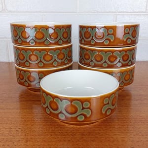 May include: A set of six ceramic bowls. The bowls are brown with a green and brown floral pattern around the rim. The interior of the bowls is white. The bowls are stacked in pairs on a wooden surface.