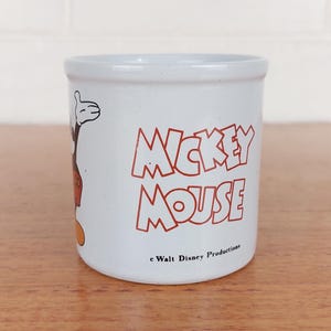 May include: A white ceramic mug with a black handle and the text "But First, Coffee" in a bold, black font. The mug is set against a blurred background, suggesting a cozy setting.