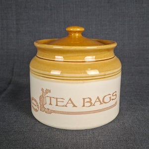 May include: A cylindrical ceramic storage jar with a removable lid, featuring a cream-colored base and a honey-brown glazed rim and top. The front displays the brown text TEA BAGS alongside a decorative illustration of a person holding a tray.
