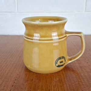 May include: A mustard yellow ceramic mug with a glossy crackle glaze finish, a flared rim, and a sturdy handle. A circular black stamp on the lower side reads Bendigo Pottery 1858 Australia, featuring a distinct horizontal ridge around the body.