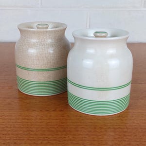 Puede incluir: Dos tarros de cerámica con tapas. Uno es beige con acabado craquelado, el otro es blanco. Ambos tienen rayas horizontales verdes alrededor de la base. Los tarros están sobre una superficie de madera.