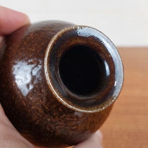May include: A small, round, brown ceramic vase with a textured surface. The vase has a dark brown rim and a wide opening. The interior is black. The vase is held in a hand.