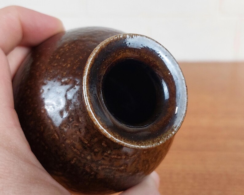 May include: A small, round, brown ceramic vase with a textured surface. The vase has a dark brown rim and a wide opening. The interior is black. The vase is held in a hand.