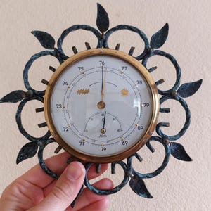 May include: An antique barometer with a brass-colored bezel and a white face. The face displays pressure readings and is surrounded by a decorative, dark teal metal frame with leaf-like accents. The barometer is held by a hand.
