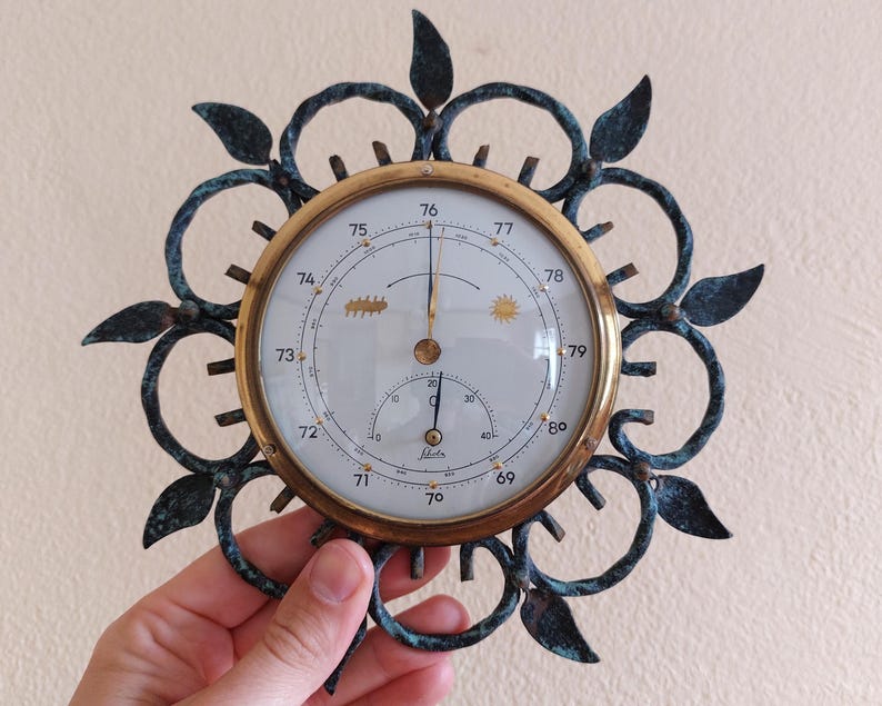 May include: An antique barometer with a brass-colored bezel and a white face. The face displays pressure readings and is surrounded by a decorative, dark teal metal frame with leaf-like accents. The barometer is held by a hand.