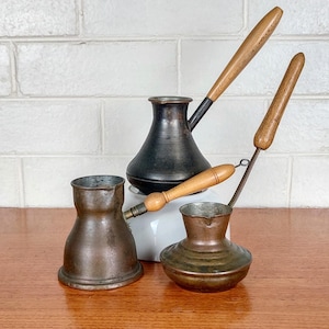 May include: Four antique copper and black metal coffee pots with wooden handles. The pots vary in size and shape, with one having a long handle and another a small, round base. They are arranged on a wooden surface against a white brick wall.