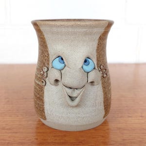 May include: A beige and brown ceramic mug featuring a sculpted face with two bright blue eyes, a prominent nose, and a wide, open mouth. The mug has a speckled texture and small circular clay accents on the sides near the eyes and cheeks.