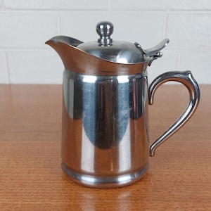 May include: A stainless steel creamer jug with a curved handle and a hinged lid. The jug has a spout for easy pouring and a small knob on top of the lid. The surface is highly reflective, showing the surrounding environment.