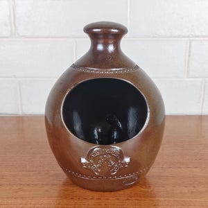 May include: A brown ceramic decorative piece with a round body and a small opening. The piece has a dark interior and a decorative emblem on the front. The piece is sitting on a wooden surface.