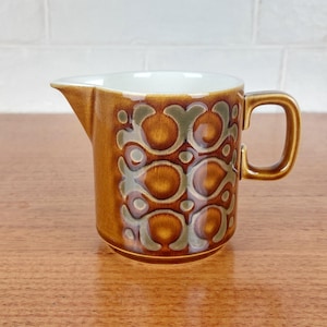 May include: A brown ceramic creamer with a decorative pattern in shades of brown and green. The creamer has a handle and a spout for easy pouring. The interior is white. This vintage piece is perfect for serving cream or milk.