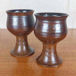 May include: Two handmade brown ceramic goblets with a dark rim and a rounded bowl shape. The goblets have a stem and a base, and a small circular emblem is visible on the stem. The goblets are on a wooden surface.