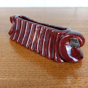 May include: A maroon ceramic vase with a ribbed design and a curved shape. The vase has a rectangular opening and a glossy finish. A small metal tag on the side reads "Made in Australia Diana Pottery."