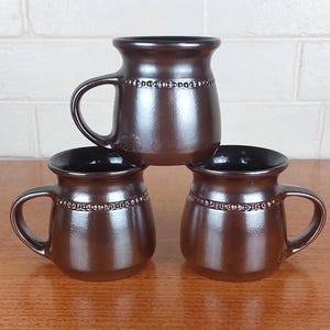 May include: Three brown ceramic mugs with black interiors are stacked on a wooden surface. Each mug has a handle and a decorative band of small, raised dots around the middle. The mugs have a glossy finish and a classic, rounded shape.
