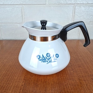 May include: A white teapot with a glossy finish featuring a blue floral pattern on the front. The teapot has a black handle, a metallic silver band around the neck, and a white lid topped with a small black knob for easy handling and pouring.