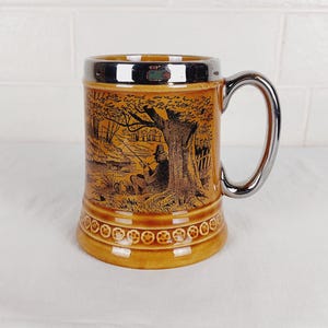 Puede incluir: Un jarro de cerveza de cerámica marrón con un acabado plateado. La taza presenta una ilustración detallada en negro y marrón de una persona pescando junto a un árbol. El asa y el borde son plateados. La base tiene un patrón decorativo.