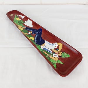 May include: A long, rectangular wooden tray with a dark red finish. The tray features a colourful painted design of a person in a white shirt, blue shorts, and a green hat, surrounded by green foliage.