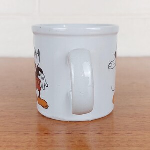 May include: White ceramic mug featuring a cartoon image of Mickey Mouse. The mug has a curved handle and a cylindrical shape. Mickey Mouse is depicted in his classic outfit with black shorts, yellow shoes, and white gloves.