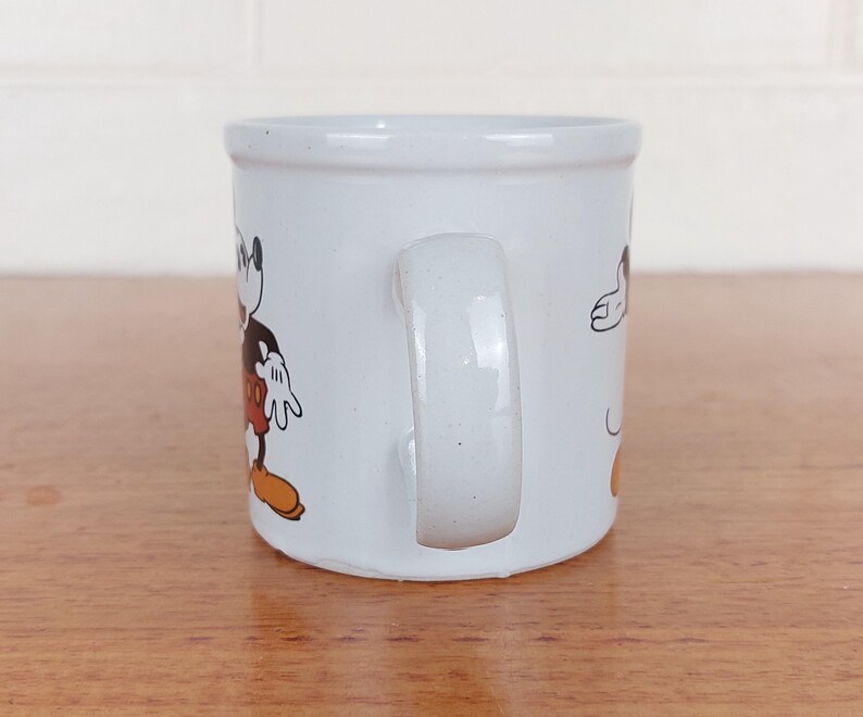 May include: White ceramic mug featuring a cartoon image of Mickey Mouse. The mug has a curved handle and a cylindrical shape. Mickey Mouse is depicted in his classic outfit with black shorts, yellow shoes, and white gloves.