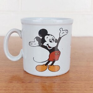 May include: A white ceramic mug featuring a classic Mickey Mouse design. Mickey is depicted in his iconic pose, wearing red shorts, yellow shoes, and black gloves. The mug has a curved handle and a speckled finish, perfect for coffee or tea.