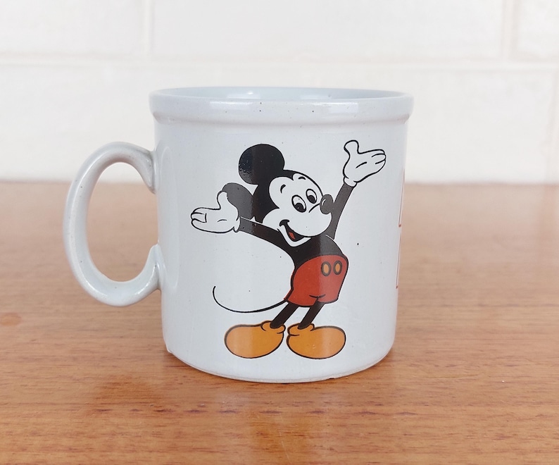 May include: A white ceramic mug featuring a classic Mickey Mouse design. Mickey is depicted in his iconic pose, wearing red shorts, yellow shoes, and black gloves. The mug has a curved handle and a speckled finish, perfect for coffee or tea.